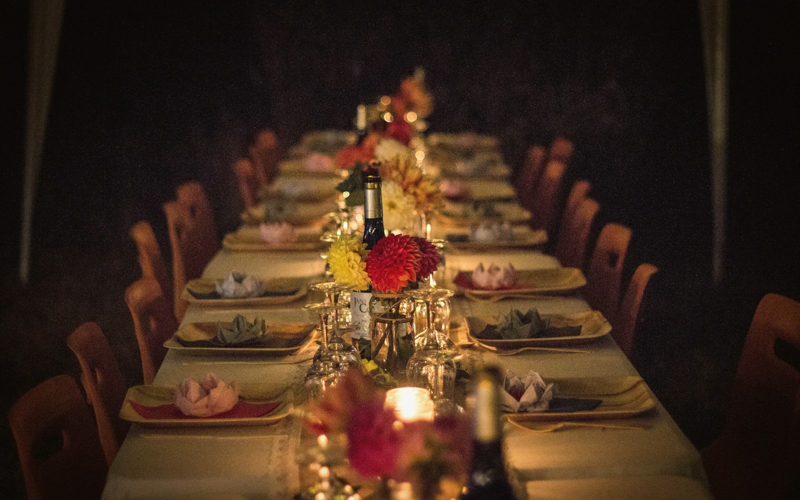 Long candlelit dinner table set with floral centerpieces, wine bottles and folded napkins