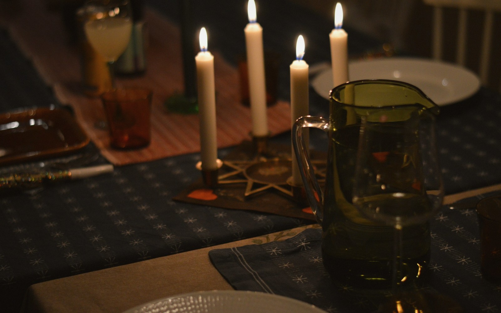 Lit candles and a glass water pitcher on a patterned tablecloth set for dinner
