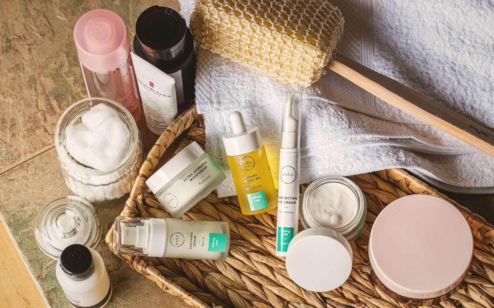 Skincare bottles, jars, and a facial brush arranged on a woven basket with towels